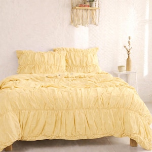 May include: A yellow bedspread set with ruffled detailing. The set includes a comforter and two pillow shams. The bedding is made of a soft fabric and is displayed on a wooden bed frame. The room has a neutral color palette.