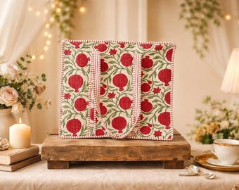 May include: A square tote bag with a pomegranate pattern in red and green on an off-white background. The bag has two handles and is displayed on a wooden stand. The scene includes a lit candle, flowers, and a lamp.