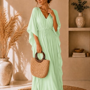 Peut inclure: Une robe caftan longue de couleur vert menthe clair avec un décolleté en V, des manches larges style kimono et une taille à cordon de serrage. La robe est accompagnée d'un sac à main en paille tressée avec deux anses rondes en bois brun foncé.
