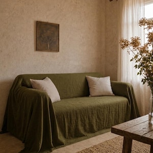 Könnte beinhalten: Ein Wohnzimmer mit einem grünen Sofa, das mit einer passenden Decke und zwei cremefarbenen Kissen bedeckt ist. Ein kleiner gewebter Teppich liegt auf dem Boden. Ein hölzerner Beistelltisch und ein gerahmtes Kunstwerk sind ebenfalls zu sehen.
