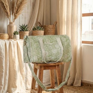 May include: A green and white floral patterned duffel bag with a shoulder strap sits on a wooden stool. The bag has a rectangular shape with a top handle and a side pocket. The background includes plants, baskets, and beige curtains.