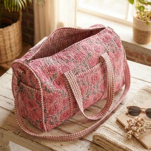 May include: Pink floral print quilted duffel bag with a matching shoulder strap and handles. The bag has a zippered top and a side pocket. The bag is sitting on a white wooden table.