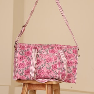 May include: Pink floral print tote bag with a matching shoulder strap. The bag features a repeating pattern of pink flowers and gray leaves on a pink background. The strap and trim are white with pink stitching. The bag is sitting on a wooden stool.