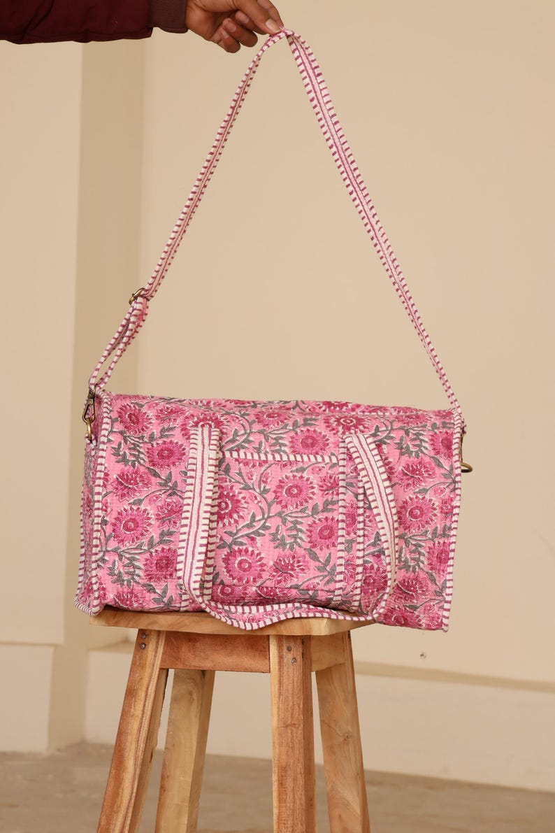May include: Pink floral print tote bag with a matching shoulder strap. The bag features a repeating pattern of pink flowers and gray leaves on a pink background. The strap and trim are white with pink stitching. The bag is sitting on a wooden stool.