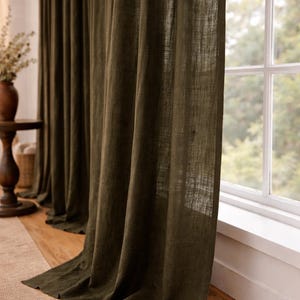 May include: Dark olive green linen curtains hanging in front of a window. The curtains are long and flowing, with a textured appearance. The window has a white frame and looks out onto a green, leafy background.