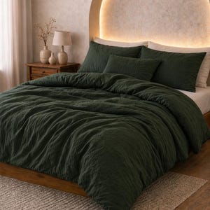 Op de afbeelding: Een donkergroene beddenset met een getextureerd dekbedovertrek en bijpassende kussenslopen. Het bed staat tegen een neutrale muur met een gebogen, verlicht hoofdbord. Een klein vloerkleed ligt aan het voeteneinde van het bed.