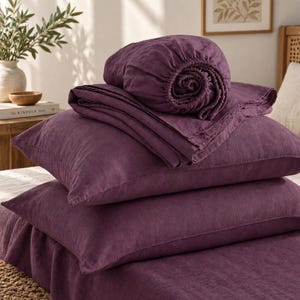 May include: A set of deep purple bedding including a fitted sheet, a flat sheet, and two pillowcases. The fabric has a visible woven texture. The items are stacked neatly, with the fitted sheet rolled at the top and the flat sheet folded below.