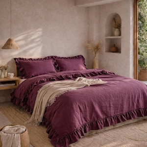 Peut inclure: Une chambre à coucher avec un lit recouvert d'une housse de couette et de taies d'oreiller violet foncé à volants. Une couverture beige est posée sur le lit. La pièce a une palette de couleurs neutres, un tapis tissé et une suspension tressée.