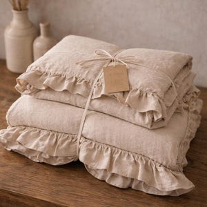 May include: A stack of beige linen bedding, including a duvet cover and pillowcases, tied with twine and a tag. The bedding features ruffled edges and is neatly folded on a wooden surface. Decorative vases are in the background.