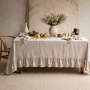 May include: A beige linen tablecloth with a ruffled edge covers a wooden table set for a meal. The table is adorned with a vase of flowers, candles, and a wooden bowl of fruit. A wooden chair is visible on the left.