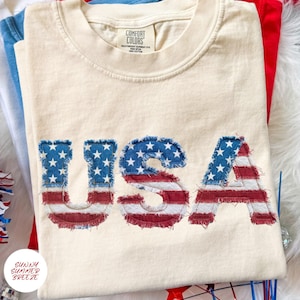 May include: A cream-colored short-sleeve t-shirt featuring the letters USA in a frayed, raw-edge applique style. The letters are patterned with a red, white, and blue American flag design. The inner neck label reads Comfort Colors.
