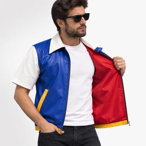 Könnte beinhalten: Eine kurzärmelige Jacke mit blauem Vorderteil, weißen Ärmeln und Kragen sowie einem leuchtend roten Innenfutter. Das Kleidungsstück verfügt über einen gelben horizontalen Akzentstreifen am unteren Saum und einen schwarzen Reißverschluss.