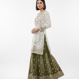 May include: A white tunic and olive green skirt ensemble with intricate gold embroidery. The long-sleeved tunic features detailed patterns around the neckline, sleeves, and front. The skirt has a flowing silhouette with matching embroidery. The outfit is a traditional style.