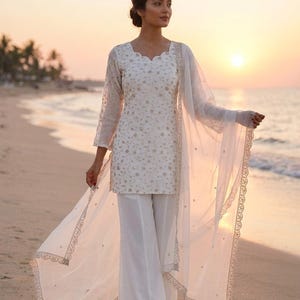 Könnte beinhalten: Weißes, besticktes Tunika- und Palazzo-Hosen-Ensemble mit einem transparenten Schal. Das Outfit hat zarte goldene Stickereien und einen wellenförmigen Ausschnitt. Geeignet für einen besonderen Anlass.