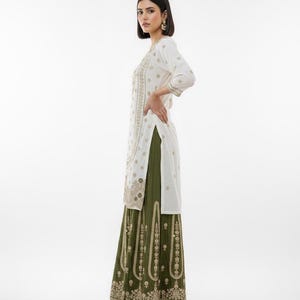 May include: A white tunic with gold embroidery and three-quarter sleeves is paired with a long olive green skirt. The outfit features detailed gold patterns and is suitable for special occasions.