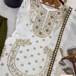 May include: White embroidered tunic and matching scarf with gold and green floral designs. The long-sleeved top features intricate embroidery around the neckline and cuffs, with a matching scarf. Olive green trousers are visible on the left.