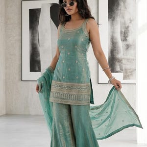 May include: A sleeveless teal green tunic with a scoop neckline and intricate gold embroidery paired with matching wide-leg trousers and a sheer dupatta. The ensemble features a metallic sheen and detailed patterns along the hem and neckline.