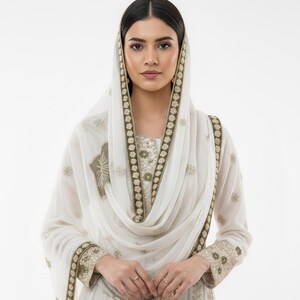May include: A white outfit, including a tunic and scarf, embellished with gold and olive green embroidery. The scarf drapes over the head. The outfit is from FIEBEE, with the logo in the bottom right corner.