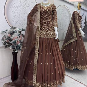 May include: A brown embroidered formal dress with a matching sheer dupatta. The dress features gold embellishments and intricate detailing. The skirt is a flowing design. A second dress is reflected in a mirror.