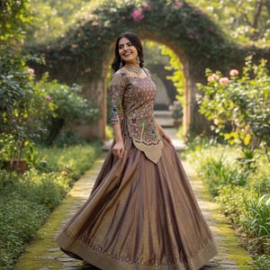 May include: A woman in a brown and gold embroidered gown and matching skirt. The gown has floral and peacock feather designs. The background shows a stone path, greenery, and a floral archway, creating a garden setting.