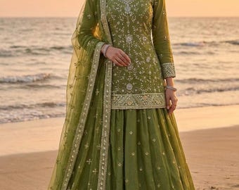 May include: An olive green embroidered formal dress with a matching dupatta. The dress has detailed silver and gold embroidery on the bodice, sleeves, and skirt. The outfit is worn on a beach with the sea in the background.