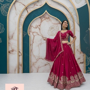 Può includere: Un completo indiano tradizionale color magenta, composto da gonna, top corto e scialle. L'ensemble è adornato da ricami dorati. Lo sfondo presenta un design marmorizzato e turchese con accenti floreali.