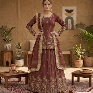 Peut inclure: Une robe indienne traditionnelle de couleur marron avec des broderies dorées. La robe comprend un haut à manches longues, une jupe et une écharpe assortie. La robe est portée avec des boucles d'oreilles dorées et un bijou de tête. La robe est présentée sur un tapis à motifs.