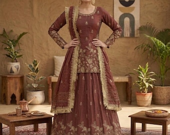 May include: A maroon Indian dress with gold embroidery. The outfit includes a long-sleeved top, a skirt, and a matching scarf. The dress is accessorized with gold earrings and a headpiece. The dress is displayed on a patterned rug.