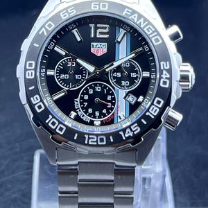 May include: A white ceramic mug with a black handle and the text "But First, Coffee" in a bold, black font. The mug is filled with a dark liquid, likely coffee, and sits on a light-colored surface.