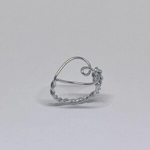 Può includere: Un anello in filo d'argento con un design a forma di cuore. L'anello presenta una fascia in filo intrecciato e un dettaglio ad anello sulla parte superiore, creando un'estetica unica e delicata. L'anello è posto su uno sfondo bianco.