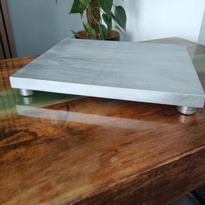 May include: A rectangular wooden platform with a light grey finish, featuring a smooth top surface and visible wood grain. The platform is elevated by four cylindrical silver-coloured metal feet positioned at each corner of the base.