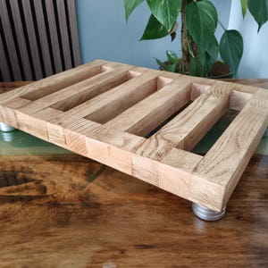 Könnte beinhalten: Ein rechteckiger Holzständer aus hellen, gemaserten Holzlatten, die mit offenen Zwischenräumen angeordnet sind. Die Konstruktion wird von vier kleinen, runden, metallisch silbernen Füßen an jeder Ecke der Basis gestützt.