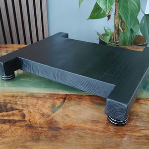 Op de afbeelding: Een rechthoekig zwart houten platform met een zichtbare houtnerftextuur, ondersteund door vier korte cilindrische voetjes. Elke voet is voorzien van drie opvallende horizontale zilverkleurige ringen rondom de omtrek.