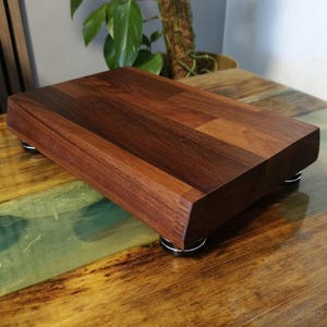 Puede incluir: Una plataforma rectangular de madera con un veteado de color marrón oscuro intenso. El bloque de madera maciza está elevado mediante cuatro pequeñas patas metálicas circulares de color plateado y negro situadas en cada esquina.