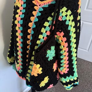May include: Black crochet poncho with a colourful granny square design in shades of yellow, orange, pink, and teal. The poncho has a V-neck and is paired with a matching black crochet bag with a long strap.