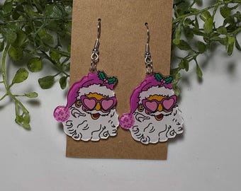 May include: A pair of dangle earrings featuring Santa Claus faces with pink hats, heart-shaped sunglasses, and white beards. The earrings are attached to silver-colored fish hook ear wires and are displayed on a brown card.