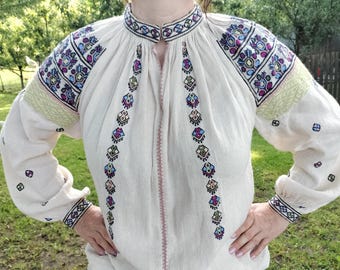 May include: Cream-colored embroidered blouse with long sleeves. The blouse features colorful floral embroidery on the shoulders, sleeves, and front. The embroidery includes shades of blue, purple, and green. The blouse has a high collar and a pink stitched seam down the front.