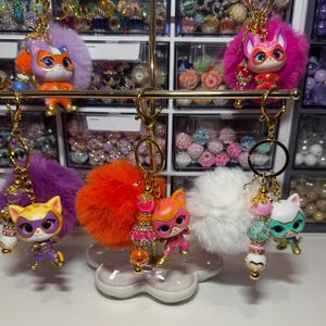 May include: Five cat-themed keychains hanging on a gold metal stand. Each keychain features a small cat figurine in a superhero mask, a fluffy pom-pom in purple, pink, orange, or white, and a strand of decorative beads with gold-tone accents.