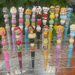May include: A collection of decorative pens with character-themed toppers. Each pen features a different character figurine, colorful beads, and a decorative base. The pens are displayed in a clear acrylic holder, showcasing a variety of colors and designs.