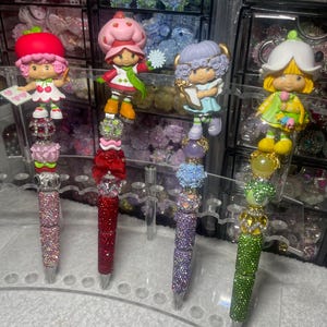 May include: A collection of decorative pens featuring colorful, sparkling designs. Each pen is topped with a different cartoon character figurine. The pens have a variety of colors, including pink, red, purple, and green, and are embellished with rhinestones.