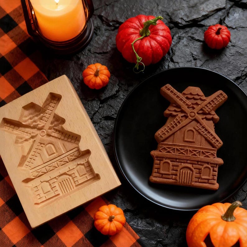 May include: A wooden windmill cookie mold and a windmill-shaped cookie on a black plate. The cookie is brown and has intricate details. Small orange pumpkins and a lit candle in a lantern are also present, suggesting a fall or Halloween theme.