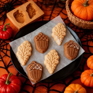 Pu&ograve; includere: Un piatto nero contiene dolcetti a forma di foglia e ghianda, alcuni spolverati di zucchero a velo. Uno stampo in legno con forme di ghianda e foglia &egrave; visibile. Zucca e un cesto con una zucca sono sullo sfondo, su una superficie arancione.