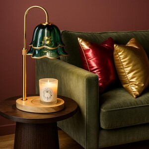 May include: A decorative table lamp with a green glass shade and gold-colored metal accents. A lit candle sits on a wooden tray below the lamp. The lamp is next to a green sofa with red and gold pillows.