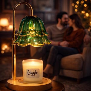 May include: A candle warmer with a green glass shade and a gold-colored metal frame. The shade is shaped like a flower. A lit candle in a glass jar sits on a wooden base. The candle jar has the word "Giggle" on it.