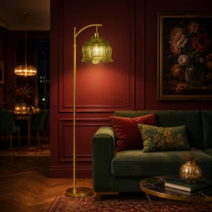 May include: A floor lamp with a slender gold-toned metal stand and a curved arm holding a translucent, olive-green glass shade shaped like a flower petal. The lamp emits a warm, soft glow, illuminating the surrounding area with a focused light.
