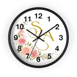 Può includere: Un orologio da parete rotondo con cornice nera. Il quadrante è bianco con numeri e lancette neri. Le lettere "SA" sono in oro. Una ghirlanda floreale di rose rosa e pesca e foglie verdi decora il lato sinistro del quadrante.