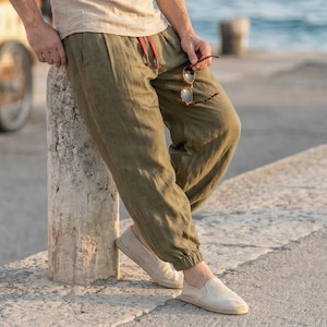 Könnte beinhalten: Olivgrüne Hose mit elastischem Bund und rotem Kordelzug, Seitentaschen sowie elastischen Beinabschlüssen. Die Hose wird mit beigefarbenen Slip-on-Espadrilles mit geflochtener Sohle und einem hellen Oberteil kombiniert.
