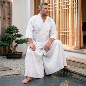 Könnte beinhalten: Ein weißer Kimono im Wickelstil und eine passende Hose mit weitem Bein. Das Outfit besteht aus einem leichten, strukturierten Stoff. Die Person sitzt in der Nähe eines Bonsai-Baums und eines Holzfensters.