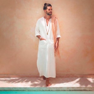 Könnte beinhalten: Ein weißes Leinen-Kaftan mit V-Ausschnitt und Vordertaschen. Ein langer, hellbrauner Schal ist über der Schulter drapiert. Eine lange Perlenkette und ein silbernes Armband sind sichtbar. Die Person steht in der Nähe eines Pools.