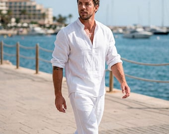 May include: A white linen shirt with a mandarin collar and long sleeves, paired with matching white linen pants. The outfit is worn by a person walking on a wooden pier near the ocean. The shirt has a buttoned placket.